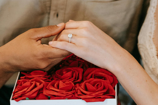 Engagement ring on a woman's hand, held by her partner, surrounded by red roses in a romantic gesture.