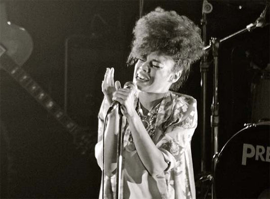 Black and white photo of a female singer performing on stage with a microphone, wearing a patterned jacket and sporting a voluminous hairstyle.