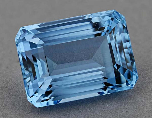 High-quality blue aquamarine gemstone with emerald cut, showcasing sharp edges and vibrant color on a neutral gray background.