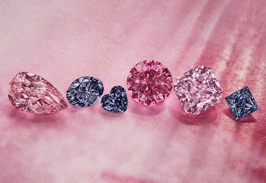 Close-up of a collection of luxury pink and blue diamonds, showcasing a variety of cuts including pear, round, heart, and square shapes. The diamonds are displayed against a soft pink background, highlighting their brilliance and elegance.