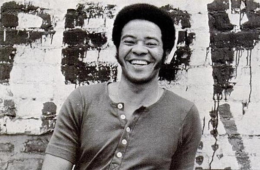 Black and white photo of smiling Bill Withers, wearing a casual shirt, standing in front of a brick wall with graffiti.