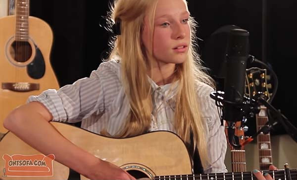Young girl playing an acoustic guitar while singing in a music studio with other guitars in the background, showcasing her musical talent.