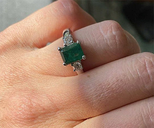 Close-up of a hand wearing a beautiful emerald engagement ring with a rectangular emerald center stone and diamond accents on either side.