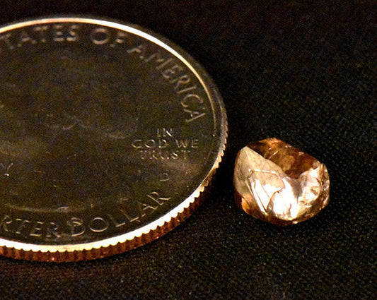 A rough gemstone next to a U.S. quarter, illustrating the size comparison of the raw, uncut gem and a coin.