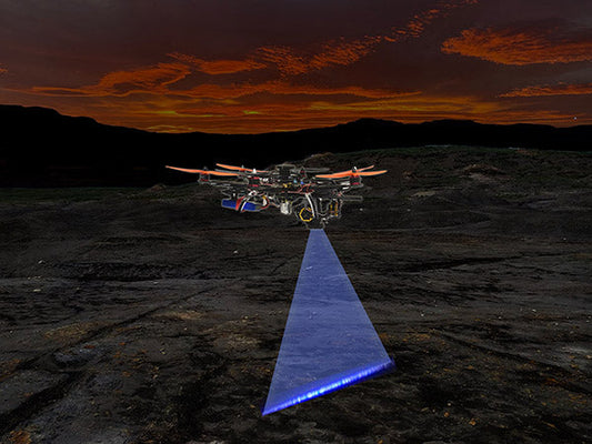 Drone flying over a rugged landscape at sunset, projecting a blue light beam downward, showcasing advanced aerial technology in an outdoor environment.