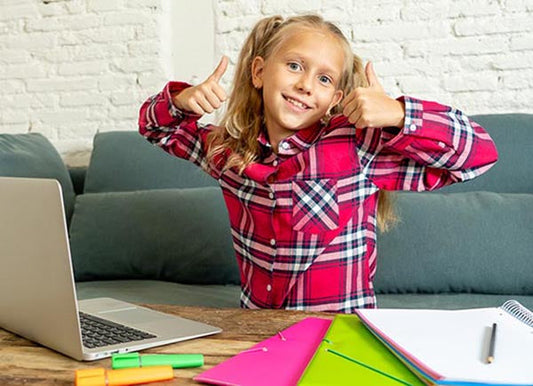 Happy young girl giving thumbs up while sitting at a desk with a laptop and school supplies, showing excitement for online learning.
