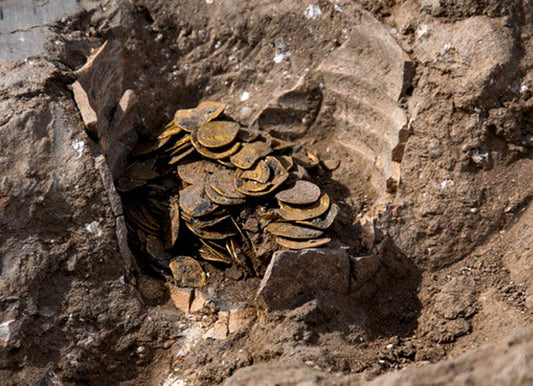 Close-up of an ancient treasure hoard of gold coins partially uncovered in the dirt. The coins are stacked together, surrounded by broken pottery shards, suggesting a historical excavation site.