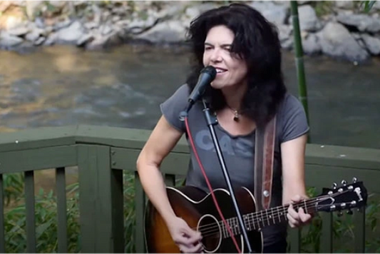 Female musician playing acoustic guitar and singing outdoors by a river.