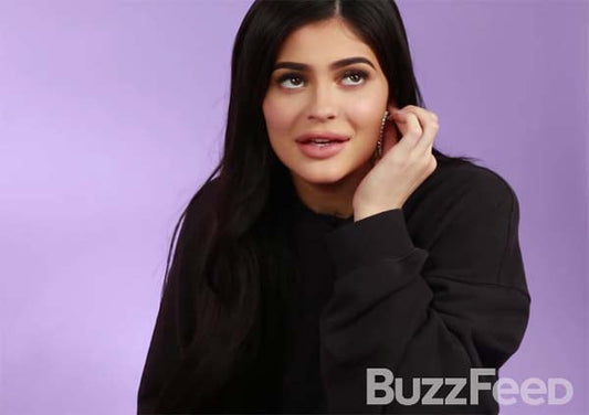 Kylie Jenner with long dark hair and wearing a black sweatshirt, looking upwards while touching her earring during a BuzzFeed interview.