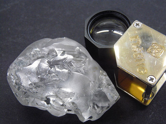 Close-up of a rough diamond crystal next to a magnifying loupe, used for inspecting gemstones. The diamond has a clear, natural appearance, and the loupe is branded with the word 'JETTING' for high-precision evaluation.