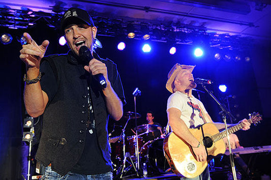 Two male musicians performing on stage, one singing with a microphone and the other playing an acoustic guitar, with a live band in the background.