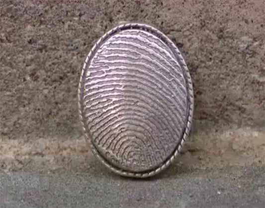 Close-up of a silver ring with a raised, textured fingerprint design, set against a simple, textured background.