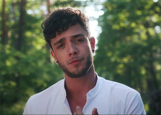 Young man with curly hair and a serious expression standing outdoors in a green forest setting, wearing a white shirt and black earrings.