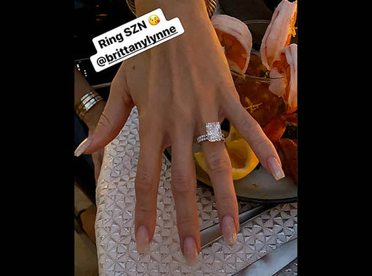Close-up of a hand displaying a stunning engagement ring with a large square diamond, accompanied by a set of complementary rings. The hand is placed next to a plate of shrimp, with a 'Ring SZN' caption visible above.