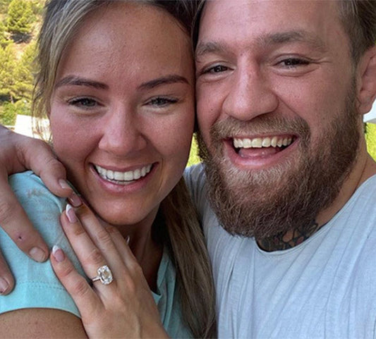 Conor McGregor and his fiancée smiling for a selfie, showing off her engagement ring with a large center diamond. The couple appears happy, posing outdoors with a lush background.