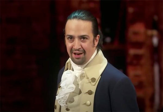 Lin-Manuel Miranda performing as Alexander Hamilton in the Broadway musical 'Hamilton', dressed in a historical costume with a white cravat and jacket, delivering an intense expression.