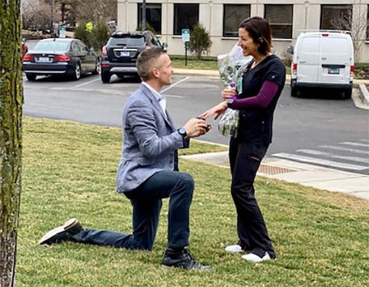 Man proposing marriage to woman in a park with a bouquet of flowers. The man is kneeling on one knee, holding an engagement ring while the woman smiles with joy in response.