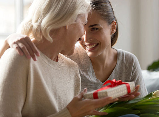 A heartwarming moment between a mother and daughter, with the daughter gifting a present wrapped with a red ribbon to her mother. The two share a joyful, affectionate smile as they enjoy their time together.