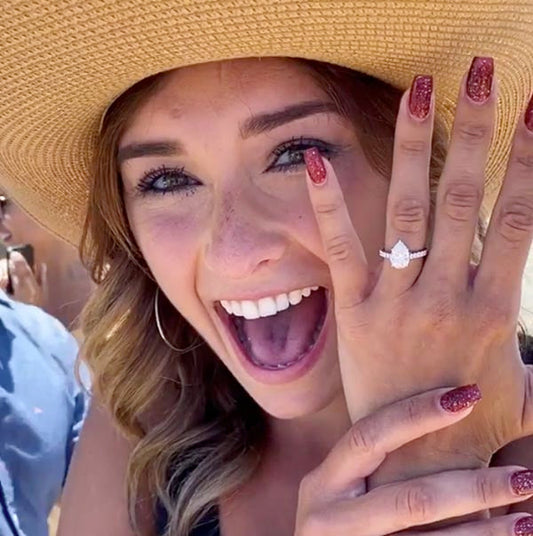 Close-up of a woman with a beaming smile, wearing a wide-brimmed hat and showing off her sparkling engagement ring with a pear-shaped diamond on her finger.