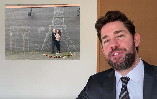 John Krasinski sitting in front of a wall with a drawing of the Eiffel Tower and a couple standing in front of it, with rose petals arranged on the ground.