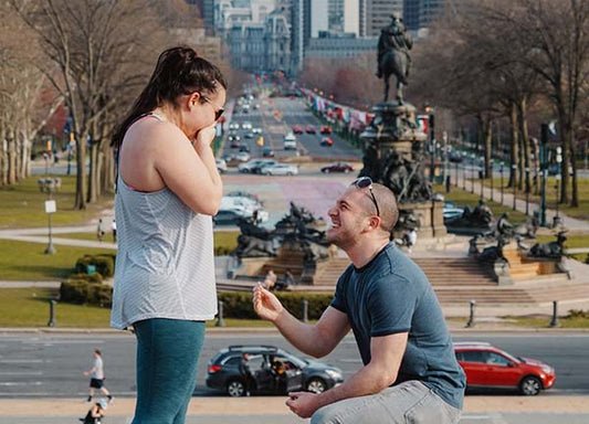 Couple engaged in a romantic proposal at the Philadelphia Museum of Art, with the man on one knee presenting a ring to his surprised fiancée.