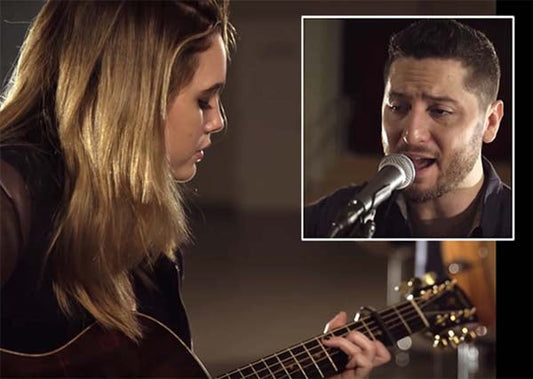 Female musician playing acoustic guitar in a recording studio while a male singer performs into a microphone in the background. Both artists are engaged in a musical duet, captured in a close-up shot of their performance.