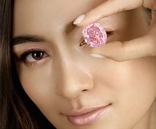 A woman holding a large pink diamond near her eye, showcasing the beauty and clarity of the gemstone.
