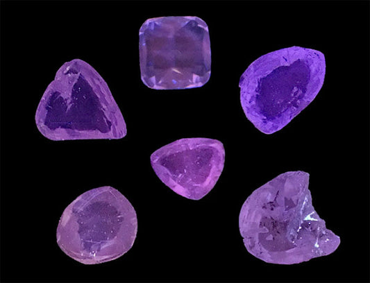 Close-up of various uncut amethyst gemstones, showcasing a range of shapes including triangular, square, and irregular forms. The stones display different shades of purple, set against a dark background to highlight their natural beauty.