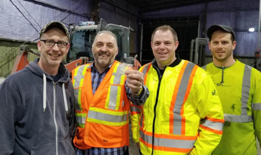 Four workers posing for a photo at a construction site, wearing safety vests and hats, with heavy equipment in the background.