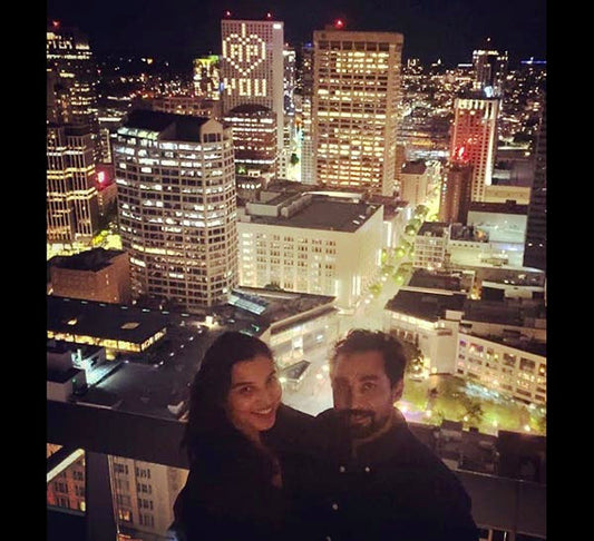 Couple posing for a photo with the San Francisco city skyline in the background at night, with a building displaying the words 'I ❤️ YOU' in lights.