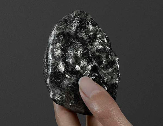 Close-up of a hand holding a dark, shiny rock with a metallic surface, possibly an uncut mineral or raw gemstone, with a grey background.
