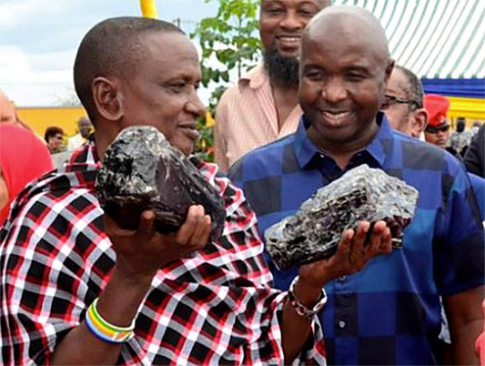 Two men, wearing traditional Maasai clothing and casual shirts, smiling while holding large pieces of raw Tanzanite gemstones during a celebration event.