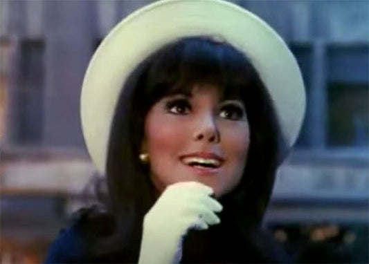 Vintage 1960s-style portrait of a woman with a white hat and gloves, smiling with a joyful expression in an urban outdoor setting.