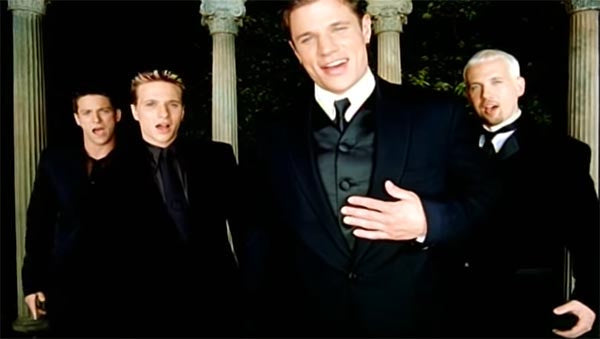Boy band performing in tuxedos with classical columns in the background, singing together on stage.