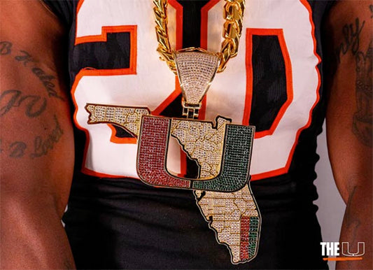 Close-up of a person wearing a large, flashy pendant shaped like the state of Florida with the 'U' logo, encrusted with diamonds and colorful gems. The pendant is part of a gold chain, and the person is dressed in a black sports jersey with the number 20.