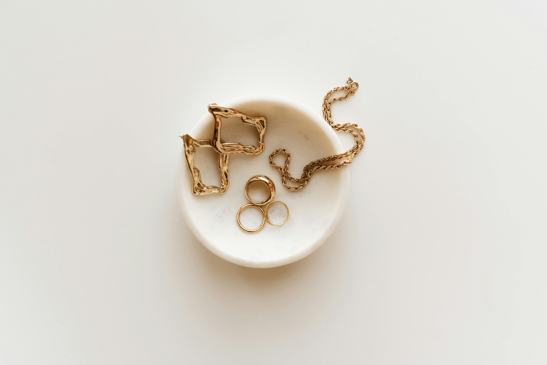 Minimalist gold jewelry including rings, earrings, and a chain displayed in a small white dish on a neutral background