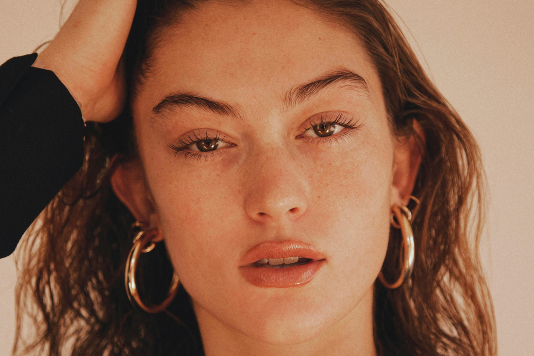 Close-up of a woman with natural makeup wearing large gold hoop earrings, her hand resting in her hair.