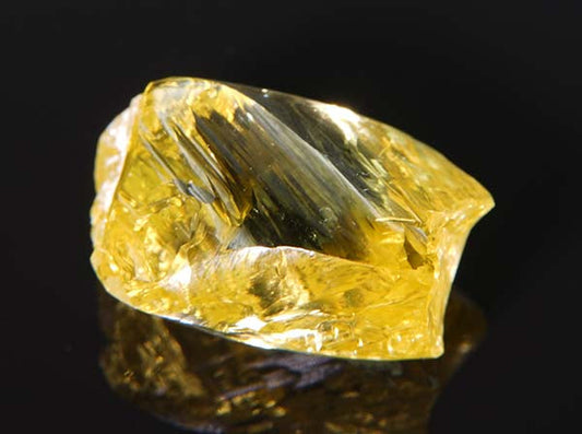 Close-up of a rough yellow diamond with visible facets, showcasing a vibrant yellow hue and reflecting light, against a dark background.