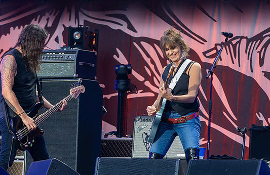 Female guitarist performing on stage with a bass player in the background, both rocking out at a live concert, with large amplifiers behind them.