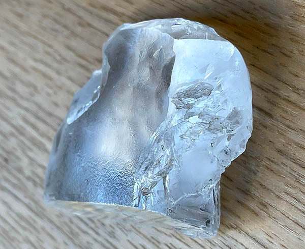Large raw crystal stone with rough edges, showing a transparent appearance, placed on a wooden surface, capturing light reflections and natural imperfections.