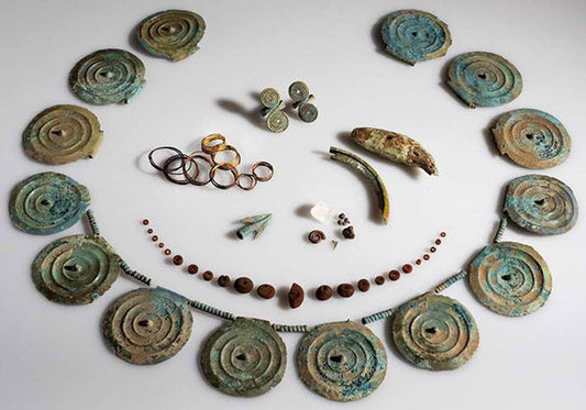 An arrangement of ancient jewelry artifacts, including circular bronze discs, rings, and beads, alongside other small objects such as shells and stones, showcasing archaeological finds.