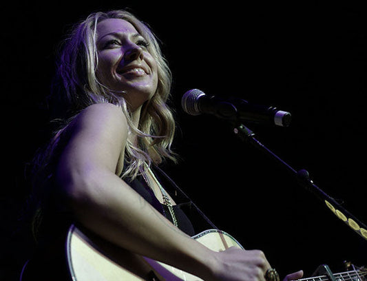 Female singer smiling while performing on stage with an acoustic guitar, microphone in front.