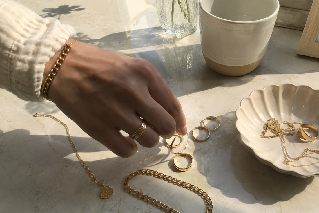 Hand arranging gold rings and delicate jewelry on a marble countertop beside a small ceramic dish.