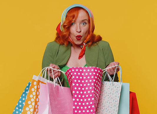 Smiling woman with red hair wearing a green jacket and colorful headband, holding multiple shopping bags in various colors and patterns against a yellow background.