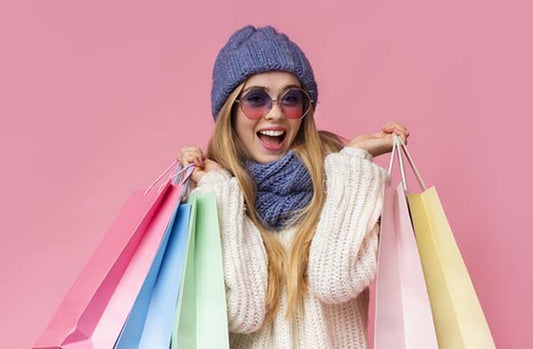 Happy young woman holding colorful shopping bags, wearing a cozy sweater, scarf, beanie, and sunglasses against a pink background.