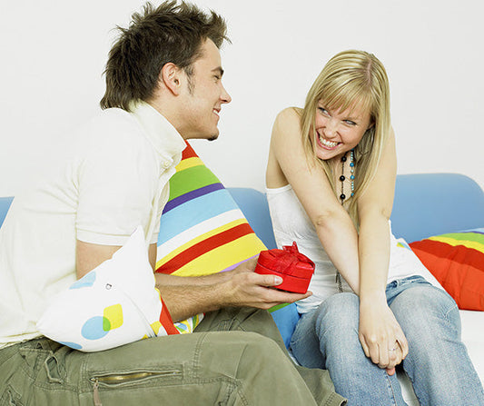A happy couple sitting together on a colorful couch with striped pillows. The man is holding a small red gift box in his hand, while the woman is smiling and excited, possibly reacting to receiving the gift. 