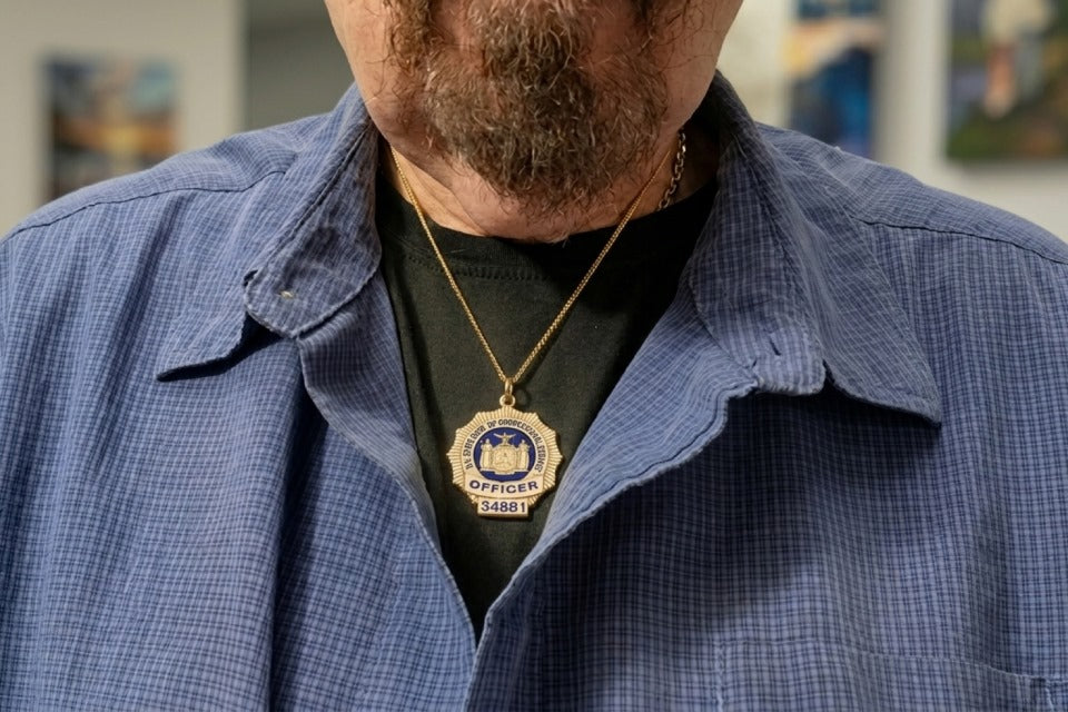 Close-up of a person’s chest and lower face, showing a gray beard and mustache. They are wearing a blue button-up shirt over a black T-shirt, and a gold chain necklace with a police badge pendant that reads