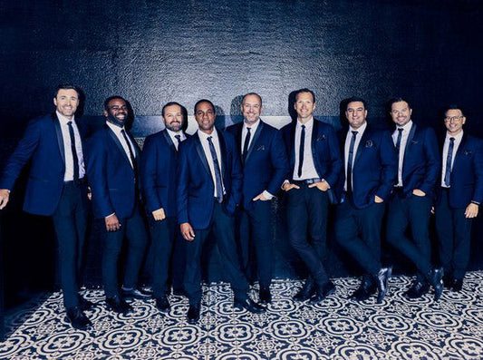 Group of nine men in formal suits posing together in front of a black wall with a patterned floor.