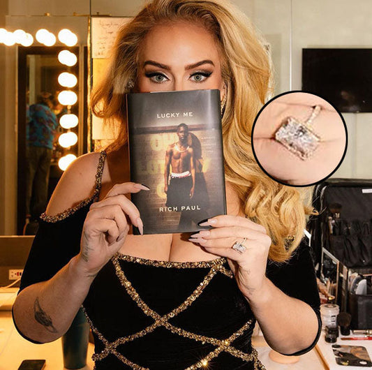 Adele holding the book 'Lucky Me' by Rich Paul, with a close-up of her engagement ring visible on her left hand. The background shows a mirror with lights in a dressing room setting.