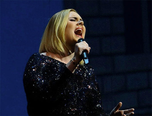 Famous singer performing on stage while passionately singing into a microphone. She is wearing a sparkly black dress and has blonde hair styled in a bob cut, with a dramatic expression as she belts out the song.
